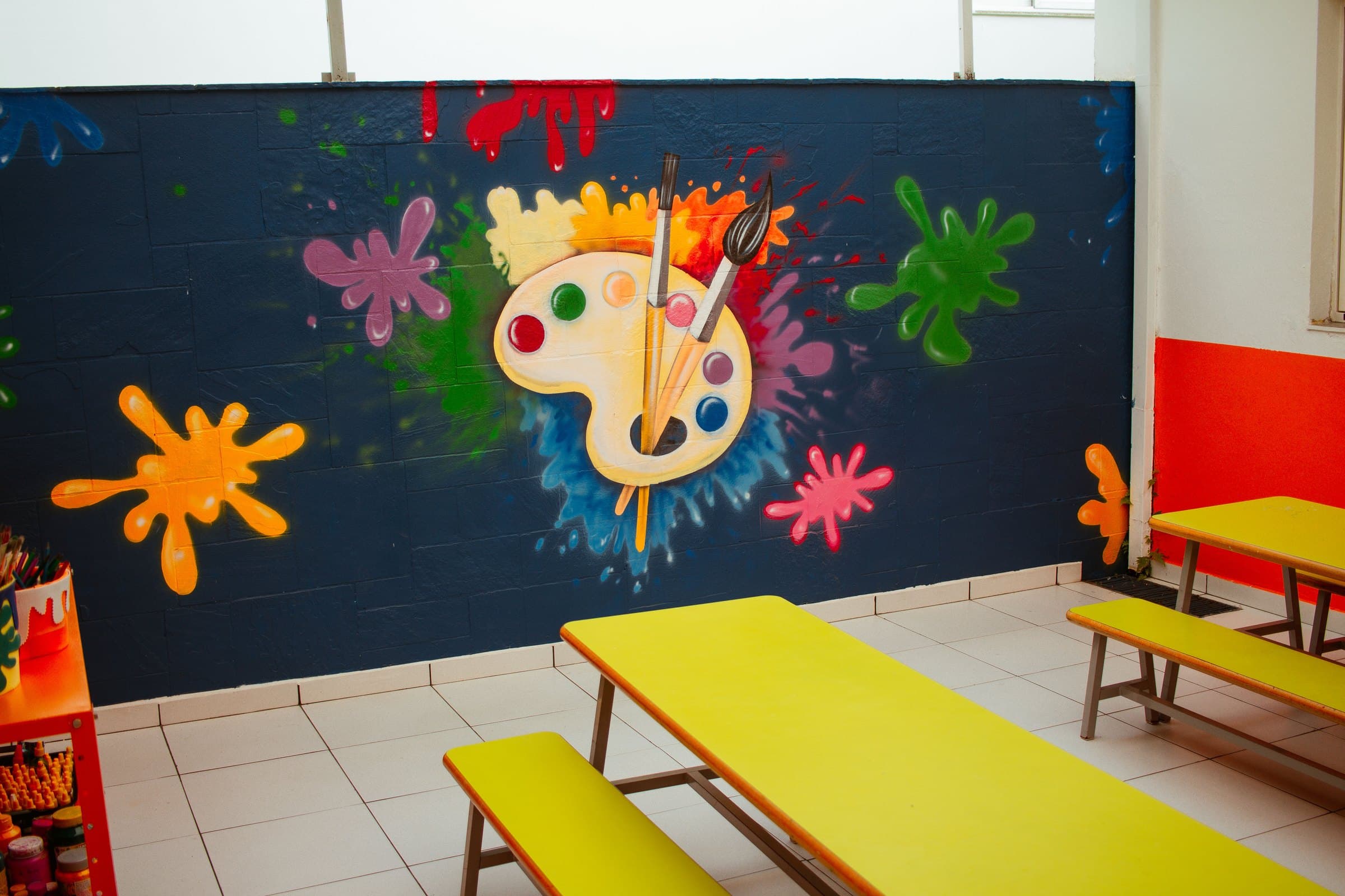 Parede pintada com uma paleta de tintas e mesas amarelas no espaço de arte do Colégio Jaguaré