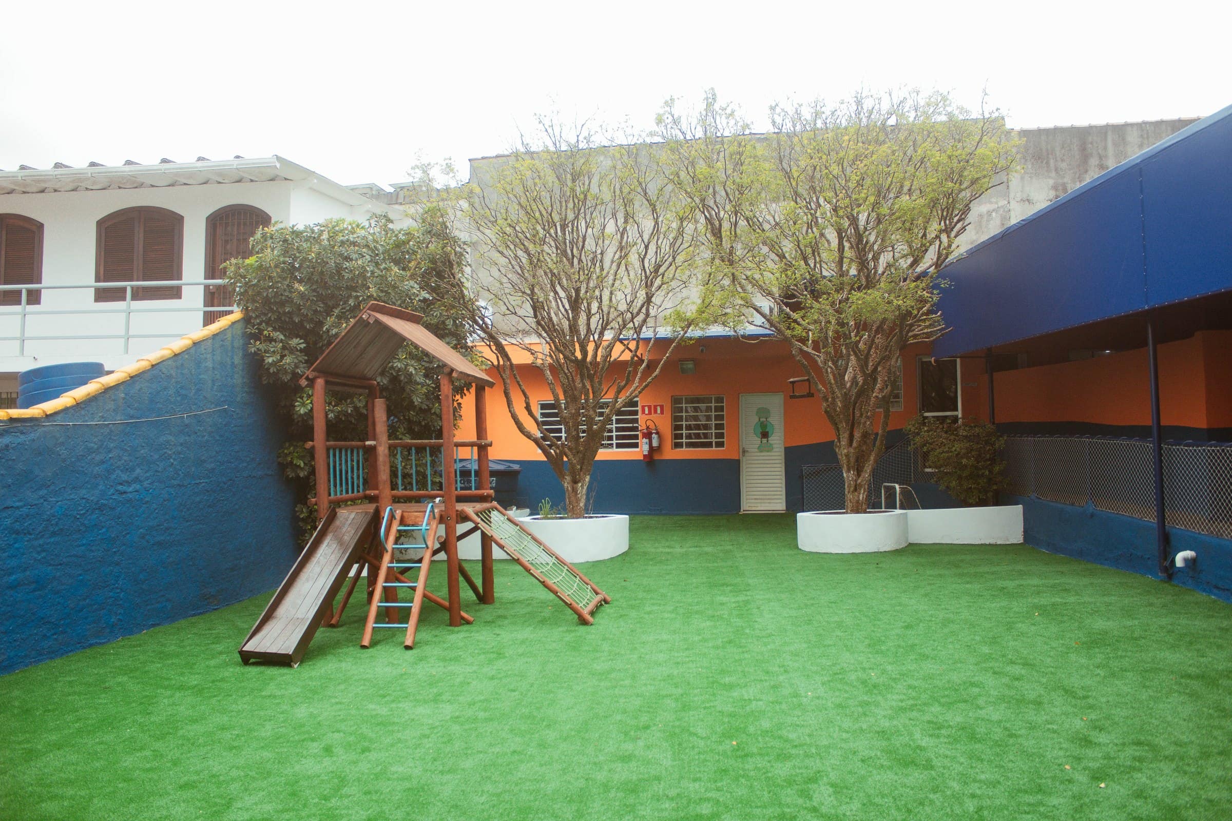 Pátio do Colégio Jaguaré com grama sintética, muro azul e brinquedos de madeira